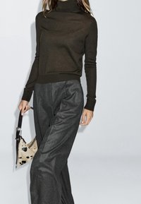 Femme portant un col roulé marron foncé et un pantalon gris à jambes larges, tenant un petit sac à main beige tacheté de noir avec une bandoulière foncée.