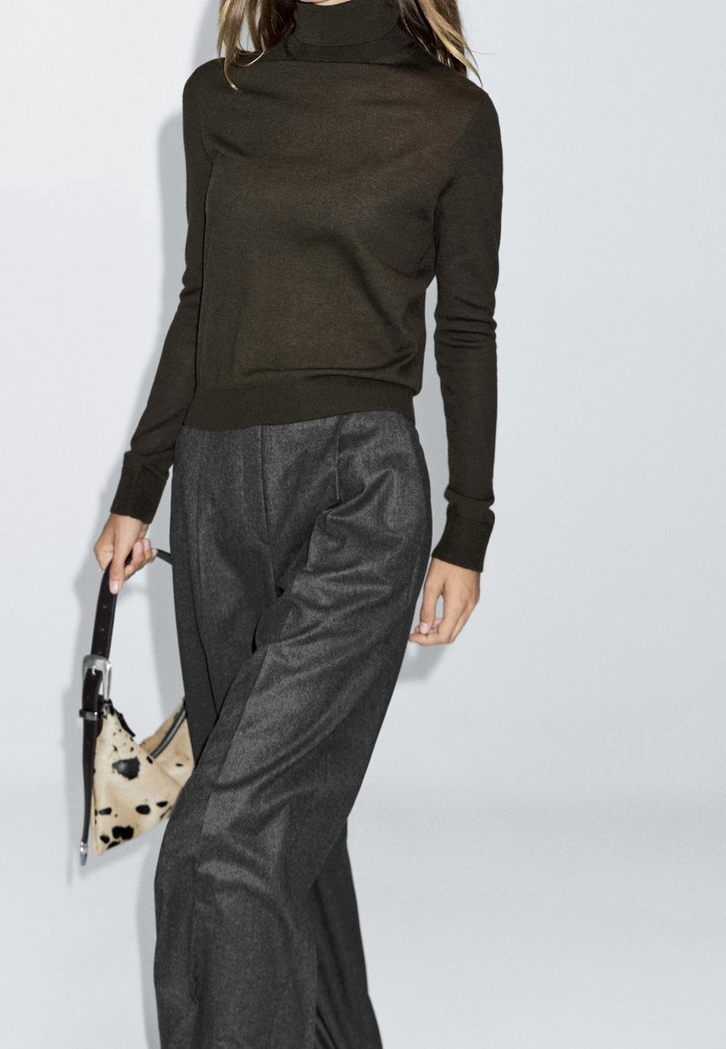 Femme portant un col roulé marron foncé et un pantalon gris à jambes larges, tenant un petit sac à main beige tacheté de noir avec une bandoulière foncée.