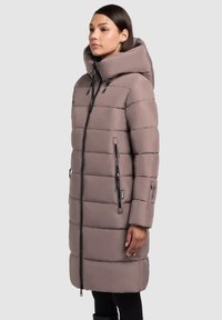 Pufferjas in zacht taupe, met een grote capuchon, verticale quiltpatronen, ritszakken en zwarte ritsaccenten. Mouwen met lange lengte.
