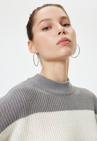 Gebreid trui met een hoge, geribbelde halslijn; heeft een verloop van grijze en crèmekleuren; textuurstof; model draagt grote oorringen.