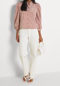 Blusa de pana rosa con mangas abullonadas y botones en la parte delantera, combinada con pantalones blancos de pierna recta y sandalias blancas con tiras. Sostiene un bolso blanco.