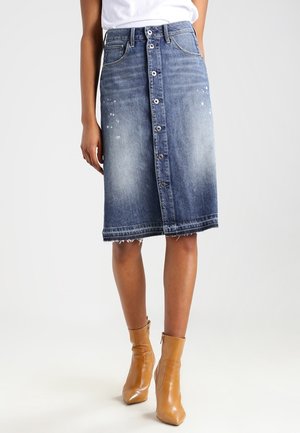 Femme portant une jupe en jean délavée mi-longue avec des boutons sur le devant, un haut blanc et des bottines beige, debout sur un fond uni.