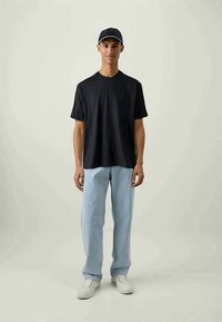 Mlad moški nosi črno kapo, črno majico, svetlo modre jeans hlače in bele superge, stoji ob preprosti svetli ozadju.