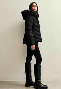 Kobieta stoi pod jasną ścianą, ubrana w czarną kurtkę puchową z futrzanym kapturem, czarne legginsy i wysokie czarne kozaki.