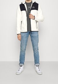 Witte fleece jas met marineblauwe schouderpanelen, contrasterende zwarte ritssluitingen, grijs t-shirt, lichtblauwe spijkerbroek en witte sneakers.