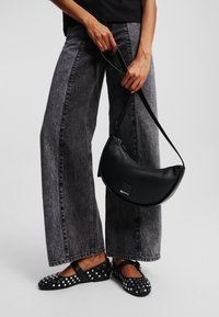 Borsa a tracolla in pelle nera con una silhouette curva e chiusura con cerniera, abbinata a pantaloni in denim grigio a gamba larga e ballerine nere con borchie.