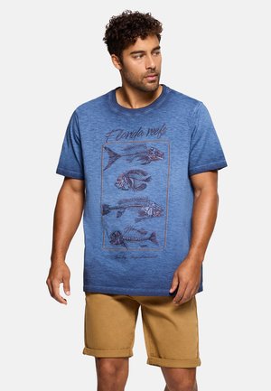 Hombre con cabello rizado que lleva una camiseta azul con el texto "Florida reefs" y esqueletos de peces, y pantalones cortos marrones enrollados, de pie contra un fondo blanco.