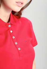 Polo rojo con cuello, con seis botones blancos y un pequeño logotipo en el lado izquierdo. La tela parece suave y texturizada.