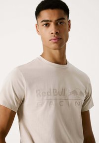 Beige t-shirt med korte ærmer; har indgraveret "Red Bull Racing"-logo; glat tekstur, klassisk crew neck-design, minimalistisk stil.
