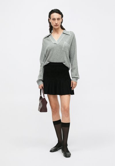 Grauer, langärmeliger Strickpullover mit V-Ausschnitt und vorderer Tasche, kombiniert mit einem schwarzen, plissierten Minirock. Die Accessoires bestehen aus einer kleinen bordeauxfarbenen Handtasche und schwarzen, knielangen Socken.