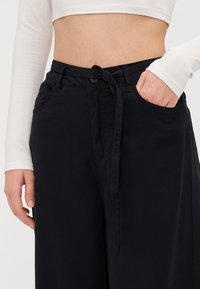Sorte vide bukser laget av myk stoff, med belte som kan knyttes, fremst lommer og knappelukking. Glatt tekstur med minimal detaljering.