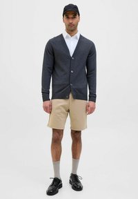 Cardigan gris foncé avec un décolleté en V et cinq boutons, associé à un short beige et des chaussures noires. Modèle portant une casquette noire et des chaussettes grises.