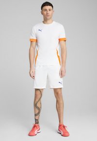 Camisa deportiva blanca con acentos naranjas y un logo azul, combinada con pantalones cortos blancos. El calzado es de color rosa brillante con suelas negras. Diseño simple.