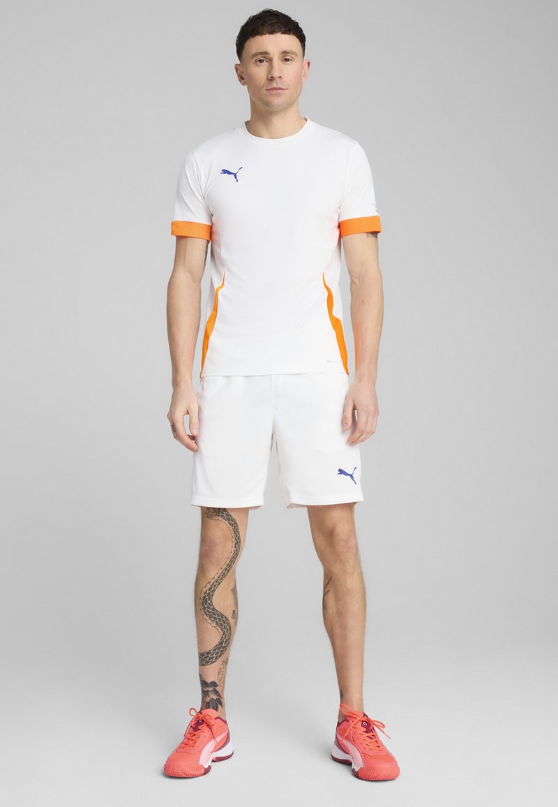 Camisa deportiva blanca con acentos naranjas y un logo azul, combinada con pantalones cortos blancos. El calzado es de color rosa brillante con suelas negras. Diseño simple.