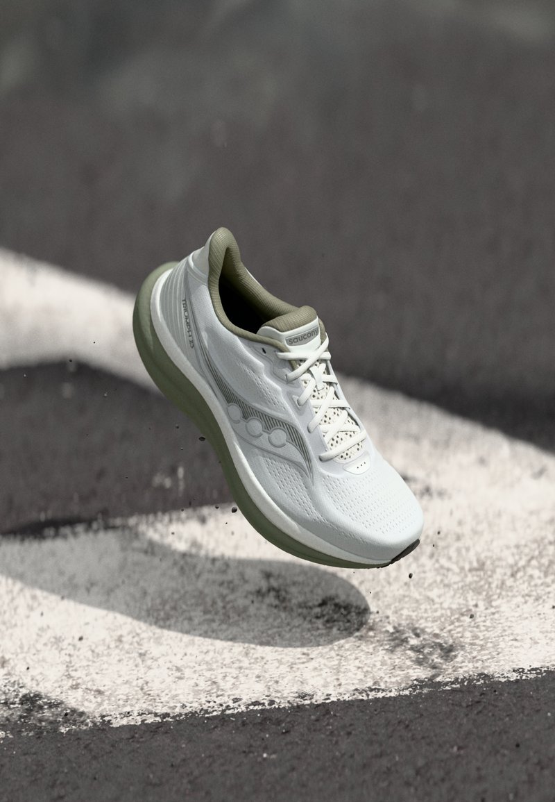 Λευκό αθλητικό παπούτσι Saucony στον αέρα πάνω από άσφαλτο με λευκές βαμμένες γραμμές, σηκώνοντας μικρά σωματίδια χαλικιού.