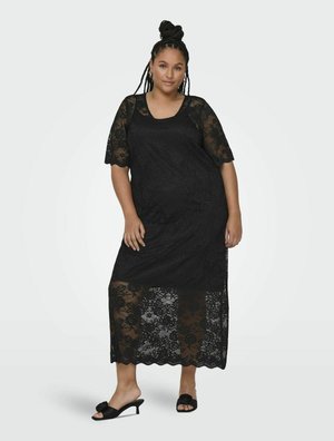 Robe maxi en dentelle noire avec des manches courtes, motif floral, décolleté arrondi et bord scallopé. Le mannequin porte des sandales noires à glissière avec un détail floral.