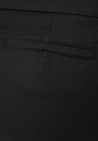 Tissu noir avec une poche passepoilée horizontale et un détail de couture verticale, probablement partie d'un vêtement comme un pantalon ou une veste.