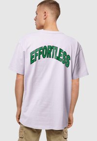 Camiseta de gran tamaño y manga corta en lavanda clara con texto verde que dice "ESFUERZO" en arco en la parte posterior. Tela de algodón, corte relajado.