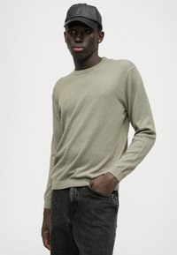 Svetlo olivno zelena pletena jopica s okroglim izrezom in dolgimi rokavi, v kombinaciji s temno sivo jean hlačami. Model pozira v sproščenem položaju.