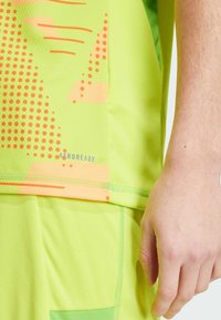 Vihreä urheilupaita, jossa on oransseja pistekuvioita ja yksityiskohtia sekä "AEROREADY" -brändäys helmoissa. Tiukka istuvuus, hengittävä materiaali.