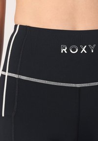 Black athletic leggings with white stitching detail, featuring a Roxy logo at the waist and a smooth, stretchable fabric texture.