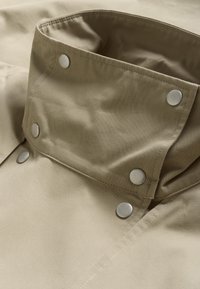 Bézs szövetkabát gallér ezüst patentgombokkal; sima felület, látható varrási részletekkel; strukturált kialakítás, hajtott gallér.