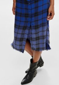 Niebieska i czarna koszulowa sukienka w kratę z guzikami, postrzępionym hemem i bocznymi rozcięciami, zestawiona z czarnymi skórzanymi botkami za kostkę.