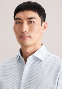 Hellblaues Hemd mit Muster und spitzem Kragen, das ein dezentes geometrisches Design und eine Knopfverschluss zeigt. Glatte Stoffstruktur.