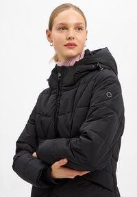 Vrouw met blond haar die gouden oorbellen met een ring draagt en een zwarte gewatteerde jas over een roze coltrui, poserend met gekruiste armen.