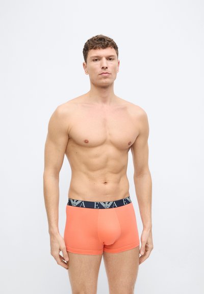 Καλογυμνασμένος νέος άνδρας που στέκεται μπροστά σε απλό φωτεινό φόντο, φορώντας κοραλλί ανδρικά εσώρουχα με μαύρη μέση με το λογότυπο "EA".