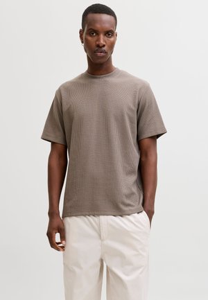 Mies seisoo, hänellä on päällään lyhythihainen savunruskea tekstuuri-T-paita ja vaaleanbeiget housut, neutraalilla taustalla.