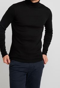 Homme portant un haut à col roulé noir ajusté et un pantalon bleu foncé devant un fond uni.