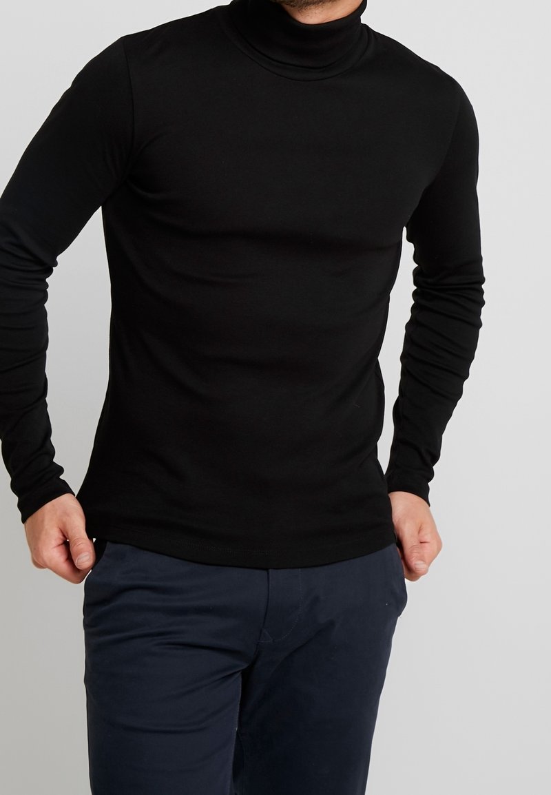 Homme portant un haut à col roulé noir ajusté et un pantalon bleu foncé devant un fond uni.