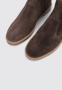 Botas de tobillo de ante marrón con paneles elásticos en los laterales, punta redonda y suela de goma marrón clara. Detalle de costura a lo largo del borde.