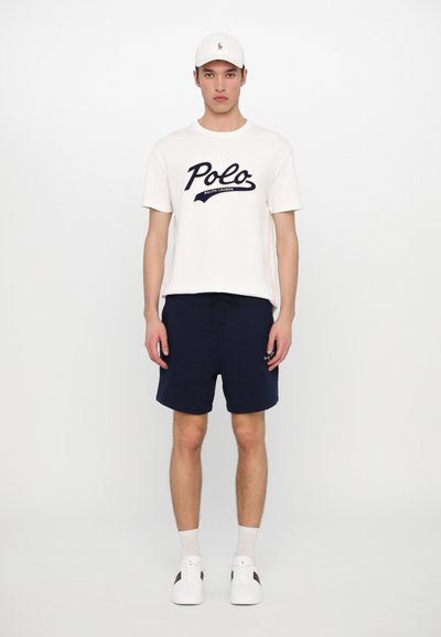 Camiseta de algodão branca com o logótipo preto "Polo Ralph Lauren", combinada com calções azul-marinho, ténis brancos e um boné branco. Design simples.