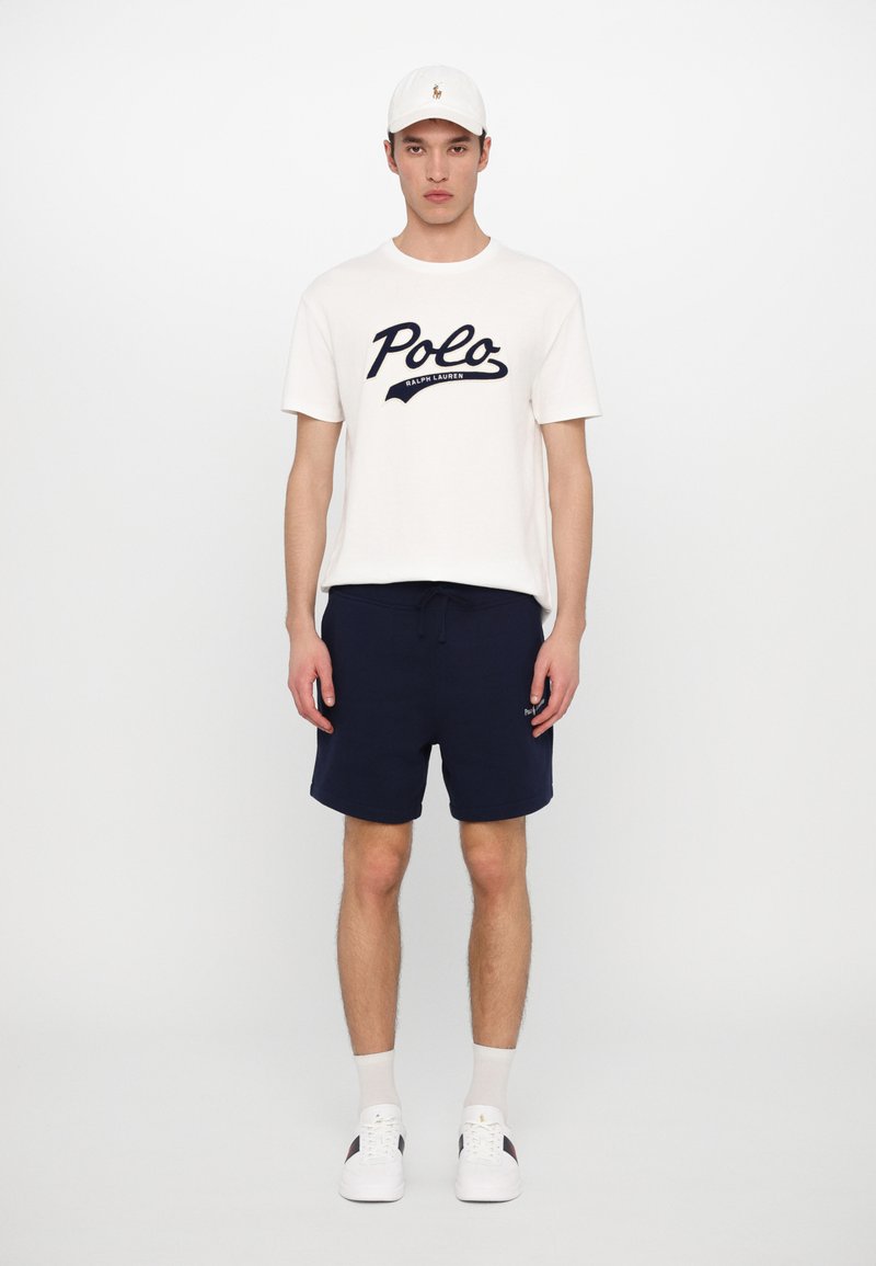 T-shirt en coton blanc avec le logo noir "Polo Ralph Lauren", associé à un short bleu marine, des baskets blanches et une casquette blanche. Design simple.