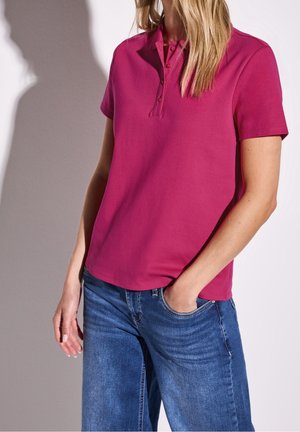 Femme portant un polo magenta à manches courtes, non rentré, sur un jean bleu, avec une main dans la poche.