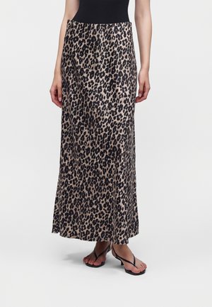 Kvinde iført langt nederdel med leopardprint, sort top og sorte sandaletter med hæl, stående foran en ensfarvet lys baggrund.
