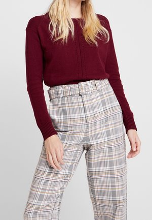 Femme portant un pull bordeaux et un pantalon taille haute à carreaux avec une ceinture assortie, debout devant un fond clair uni.