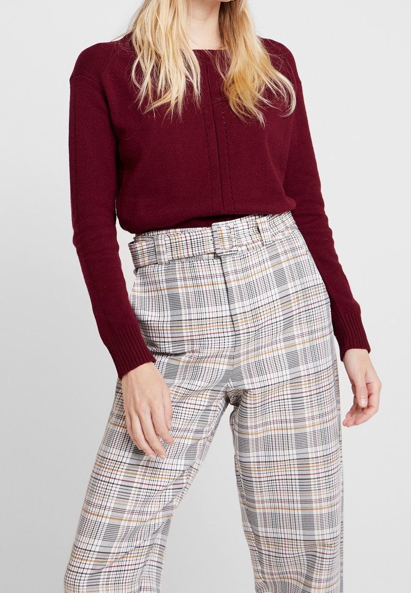 Femme portant un pull bordeaux et un pantalon taille haute à carreaux avec une ceinture assortie, debout devant un fond clair uni.