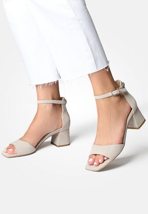 Sandali con tacco alto beige in suede opaco, con cinturino alla caviglia, punta aperta e design con tacco a blocco, caratterizzati da una texture liscia.