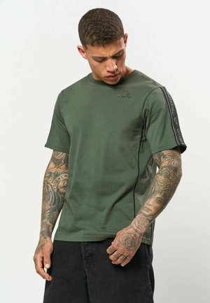 Mand med fuldærmet tatoveringer iført grøn t-shirt med sort stribe på ærmet og sorte bukser, ser ned på sin hånd.