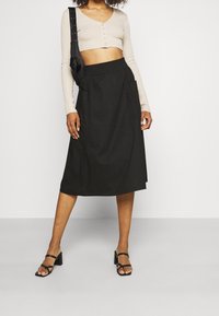 Falda midi negra con cinturilla elástica, que presenta bolsillos laterales y una textura suave. Combinada con una blusa de manga larga beige y recortada, y unos tacones negros.