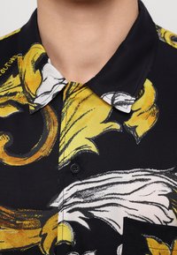 Gros plan d’un col de chemise noir avec des motifs floraux abstraits jaunes et blancs et un bouton visible sur le tissu sous le col.