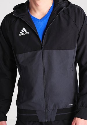 Homme portant une veste zippée Adidas noire et gris foncé par-dessus un t-shirt bleu à col en V, debout devant un fond blanc.