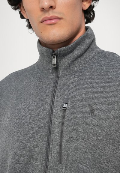 Pelēks zip-up džemperis ar teksturētu adījumu, piedāvājot augstu apkakli un smalku zīmola identifikāciju. Iekļauts rāvējslēdzēja vilkšanas elements ar logo detaļu.