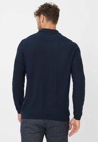 Polo a maniche lunghe di colore blu navy con tessuto testurizzato. Presenta un colletto leggero e polsini a coste, visto da dietro, abbinato a pantaloni a fantasia.