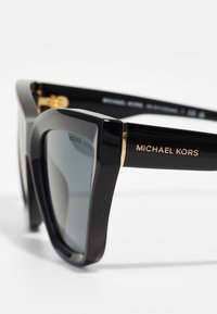 Mustat, suuret aurinkolasit kiiltävällä pinnalla, kultaiset yksityiskohdat saranoiden lähellä ja "MICHAEL KORS" -logot temppeleissä.