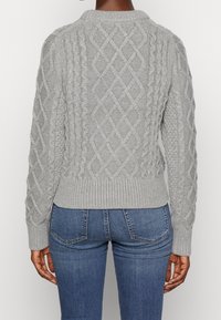 Tylny widok osoby w szarym swetrze w warkocze i niebieskich dżinsach, stojącej na tle białego tła.