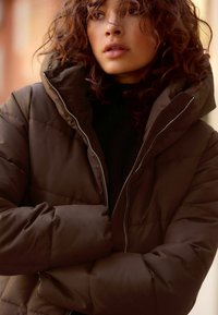 Veste en duvet marron avec un col haut, un design matelassé et des détails de fermeture éclair en argent, présentant une texture douce et lisse ainsi qu'une coupe oversized.